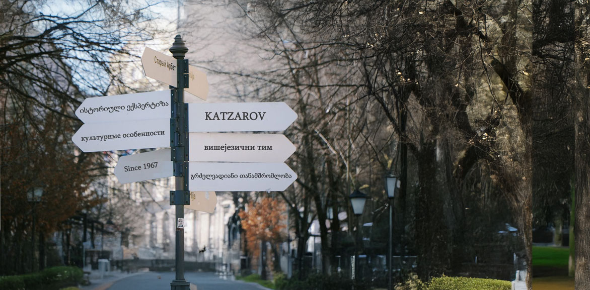 A directional signpost in a tree-lined park, featuring multiple language signs, including one pointing towards Katzarov, symbolizing multilingual expertise and global reach.